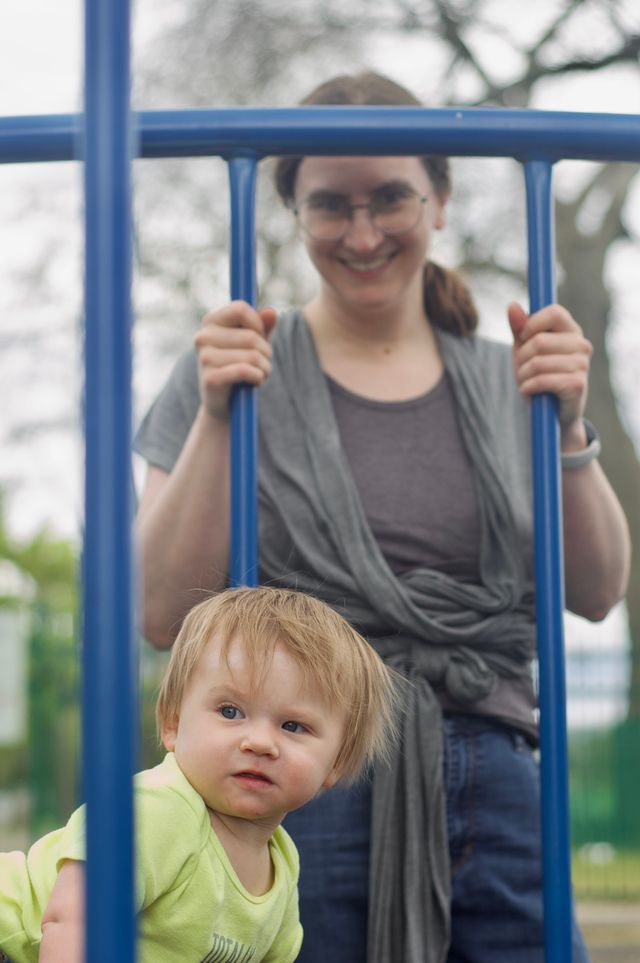 A small blonde child with a focused expression on her face crawling between two wide-spaced blue fences. In the background, behind the fence, is her mother, a smiling brunette woman looking at the camera.