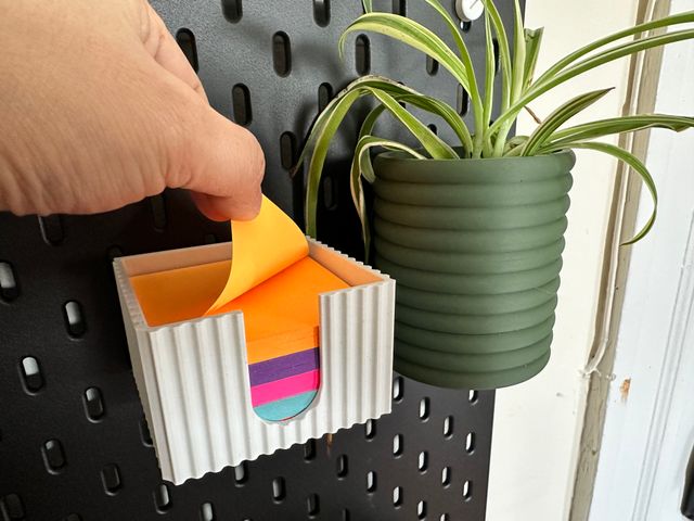 A white box with a vertically ribbed textured exterior, an open top, and a thumb slit running down the front. The box is mounted on an Ikea pegboard, next to a green plastic planter with a spider plant growing from it. Through the thumb slit a stack of multicolor sticky notes is visible. A hand is folding back the topmost note to tear it off.
