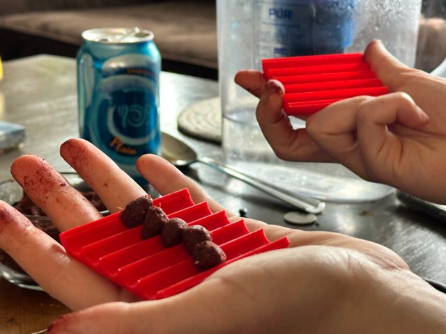 A person holding two red plastic pieces with channels running through them, one in each hand. In one of the pieces a lumpy bit of dark reddish clay is in each channel.