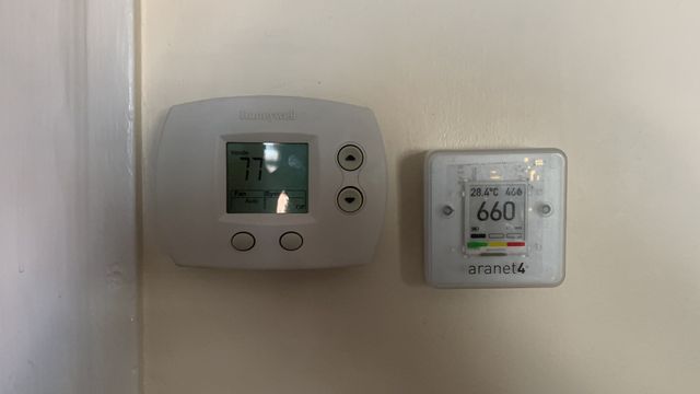 A thermostat and an Aranet4 hanging side by side on a wall. The Aranet4 is smaller than the thermostat by maybe 20%.