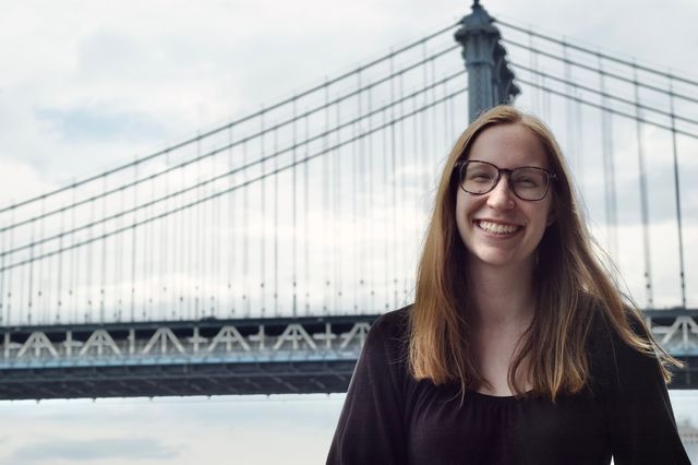 A young blonde woman in glasses and a black shirt with a greenish suspension bridge behind her.