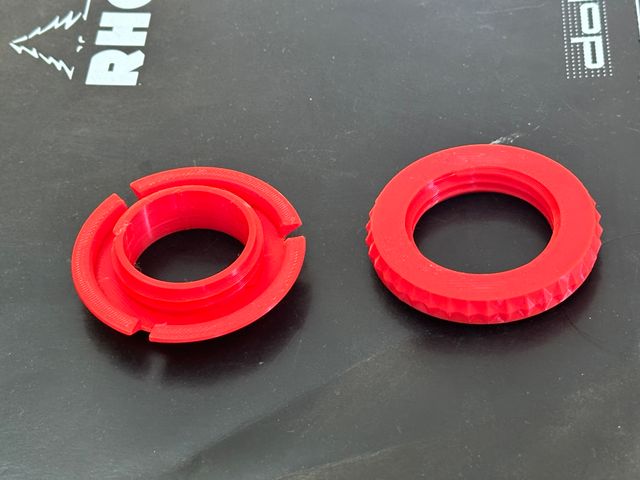 Two red plastic donut shaped objects side by side. One has three channels in the outer wall and an internal post with screw threads. The other has a knurled outer wall and internal screw threads.