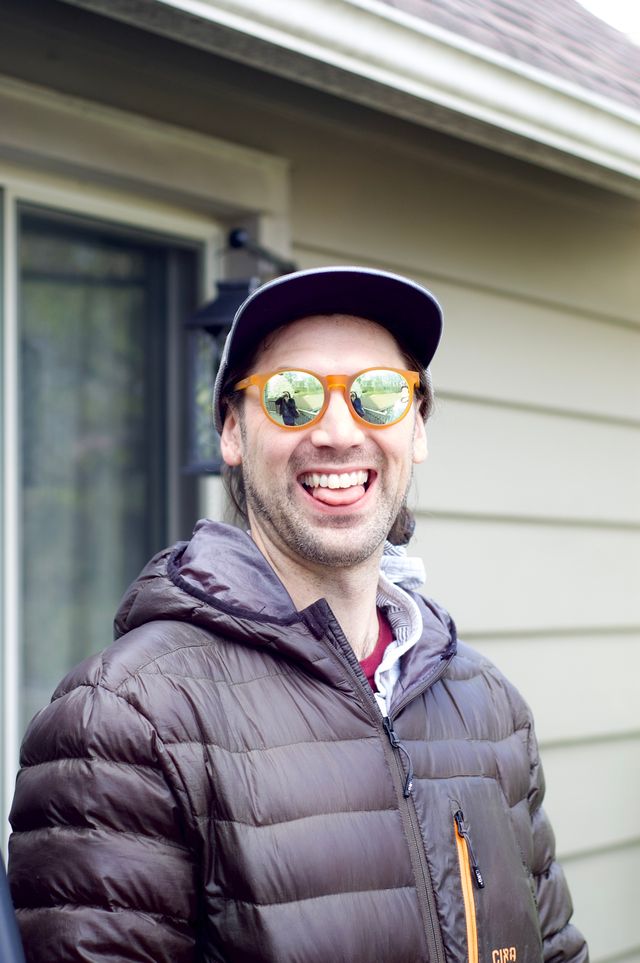 A close up of a 30-something white man wearing round orange sunglasses, a dark puffy jacket, and a cap with the brim tilted upward. He's smiling and his tongue is sticking out slightly, cheekily. In his sunglasses the reflection of the photographer can be seen, with a camera obscuring his face.