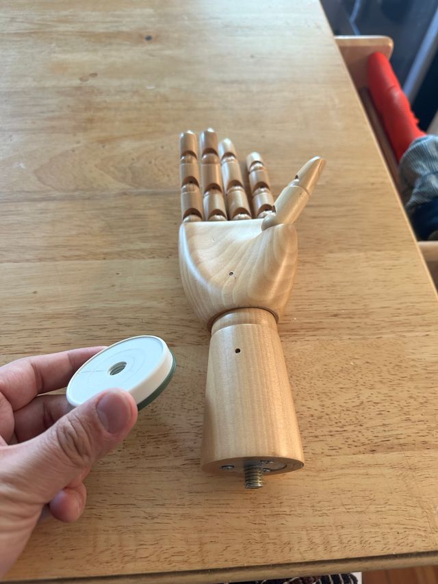 A wooden articulated hand (like for drawing) laying on a counter. There is a metal threaded rod extending from the flat base of it, below the wrist. On the left side of the hand is a (real) hand holding a green and white plastic cylinder with a hole in the middle.