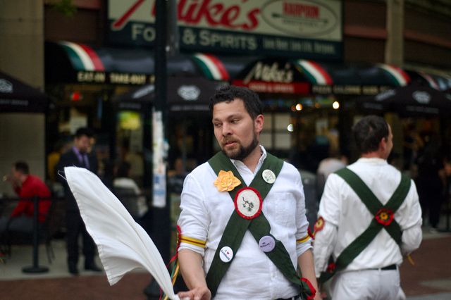 A white bearded man is morris dancing in kit. He's wearing a white shirt with red and yellow ribbons around his elbows. He has a green felt X across his chest with patches and buttons on it. He's mid-action, a white hankie in his hand arcing upward into the air. Behind him, back to us, is another man in similar garb, and behind them an out-of-focus city block.