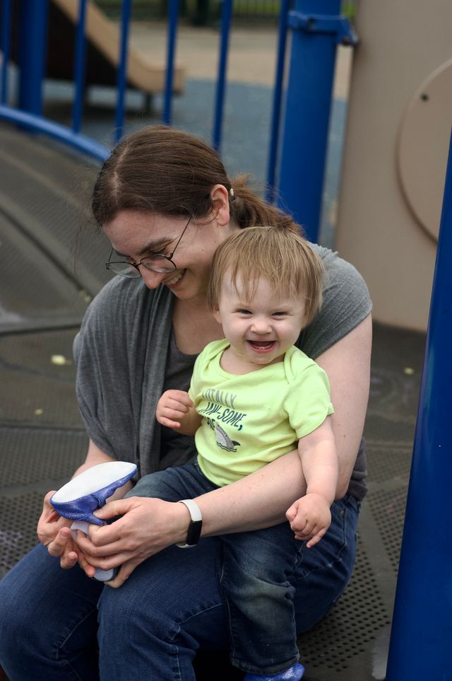 A smiling brunette white woman with glasses in her early 30s holding a small grinning blonde child who has only two lower teeth in the center of her mouth. The woman is putting a blue shoe on the child. Behind them is the blue and brown structure of a playground.