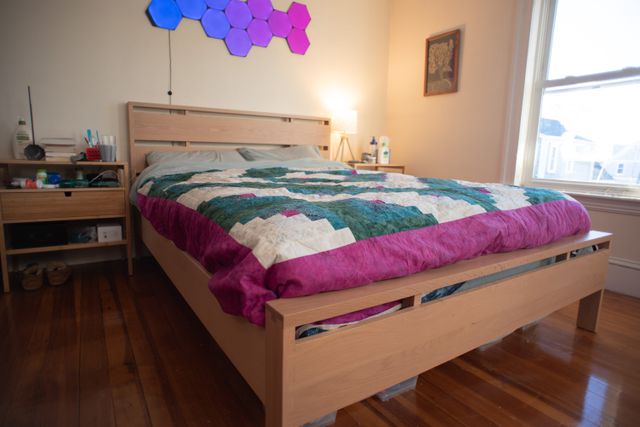 A bedroom with a light wood bed frame made of simple modern right angles and double dovetail joints at the corners. Over the mattress is a beautiful teal, pink, and white quilt with a radiating diamond pattern.