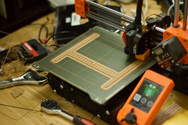 A tan capital-H shaped object mid-print. There's a broad flat tan "brim" around the base of the print.