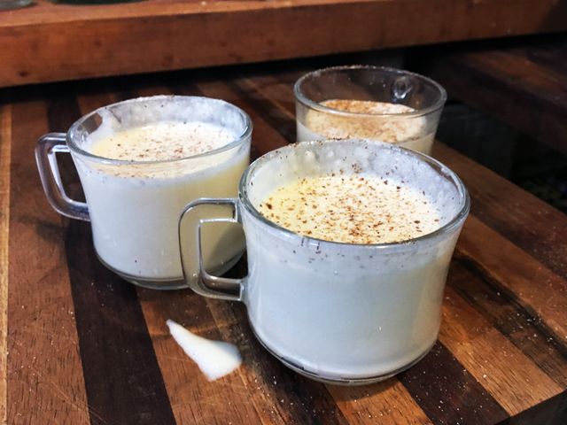Three glass mugs of thick, creamy, lightly colored egg nog with a sprinkling of nutmeg on top.